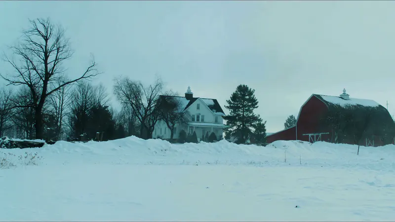 A white two storey farmhouse and a red barn in deep snow under a pale winter sky, bare trees and a pine between them