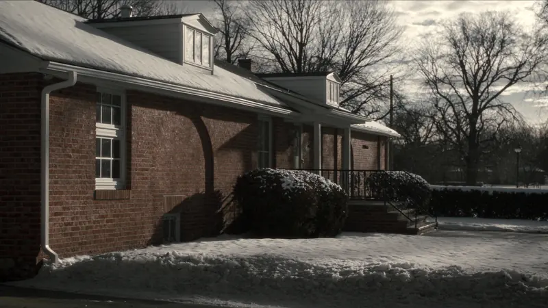 A brick ranch with dormers after snow on an overcast morning, a small basement window at the foundation, bare trees behind