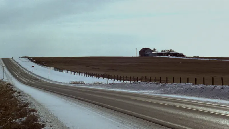 A two lane road through snowy farm fields under an overcast sky, fence posts along the shoulder and a farmstead on the horizon