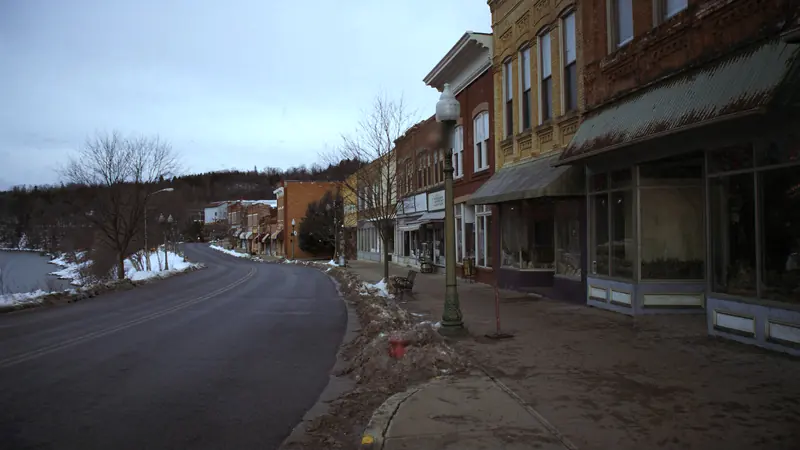 A small town main street beside a river on an overcast winter morning, old brick storefronts, a lamp post and an empty road